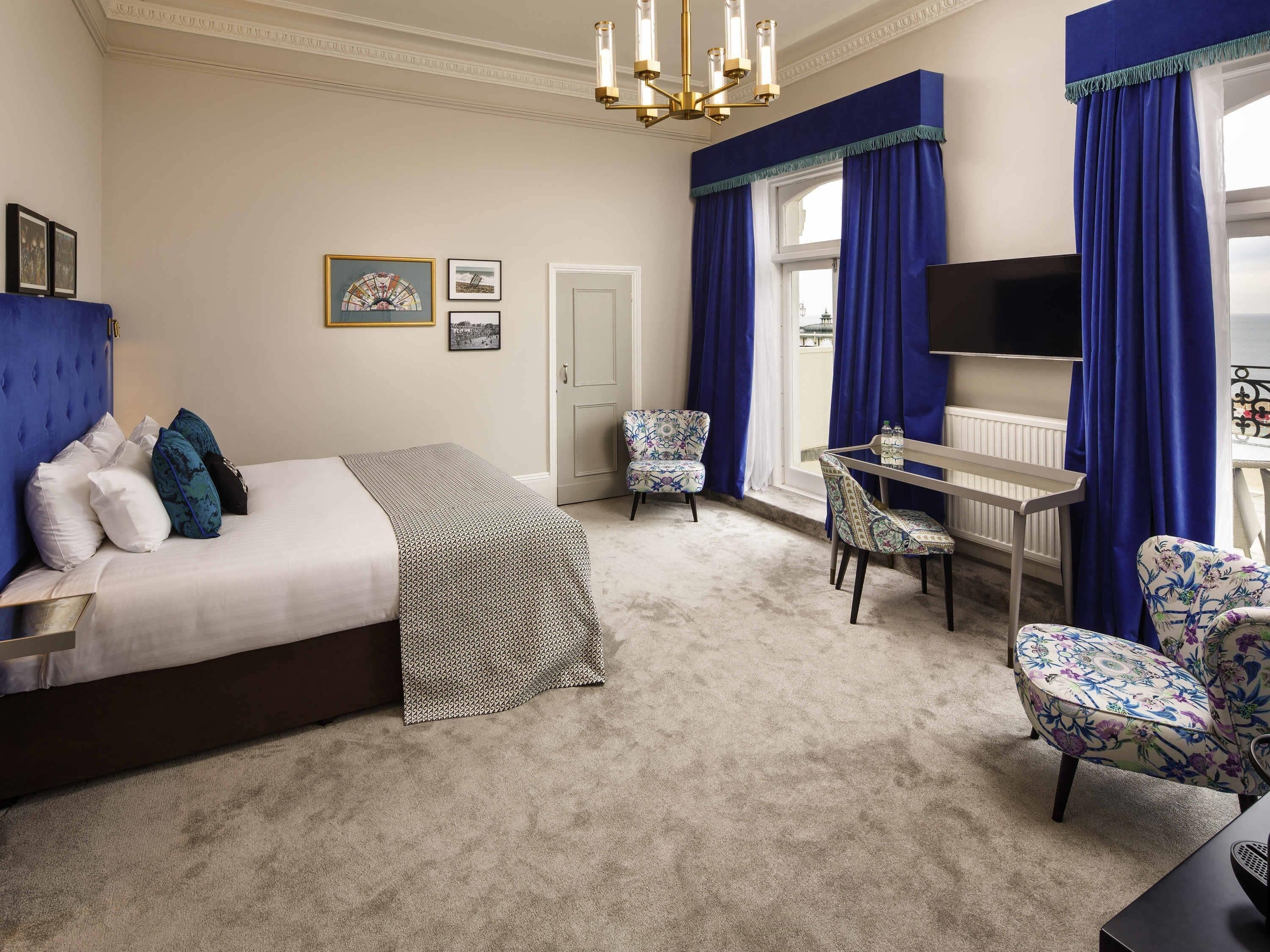 A bedroom with a bed, a desk, two chairs, a TV, and a window with curtains.