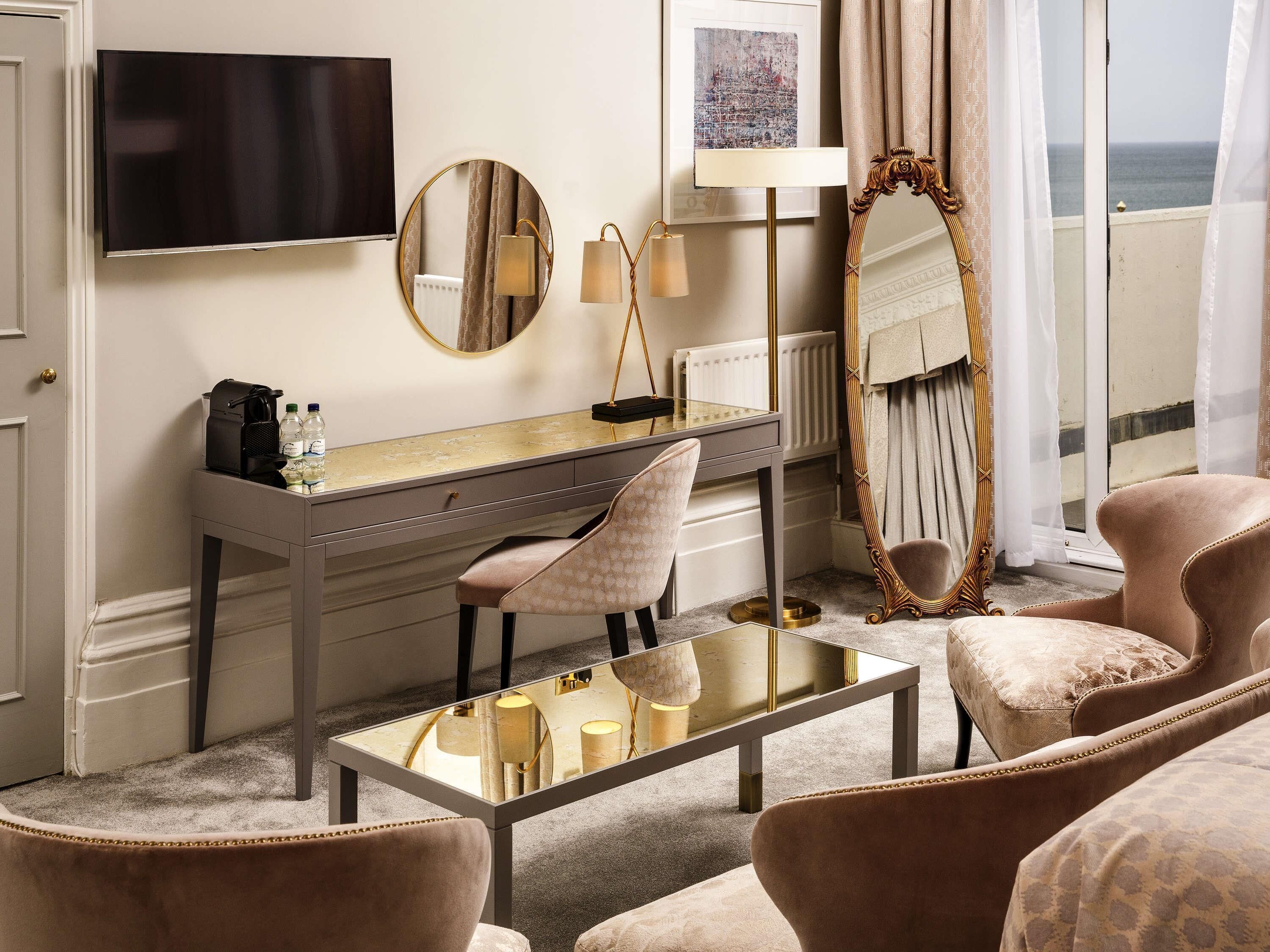A hotel room with a desk, two chairs, a large mirror, a television, and a view of the sea.