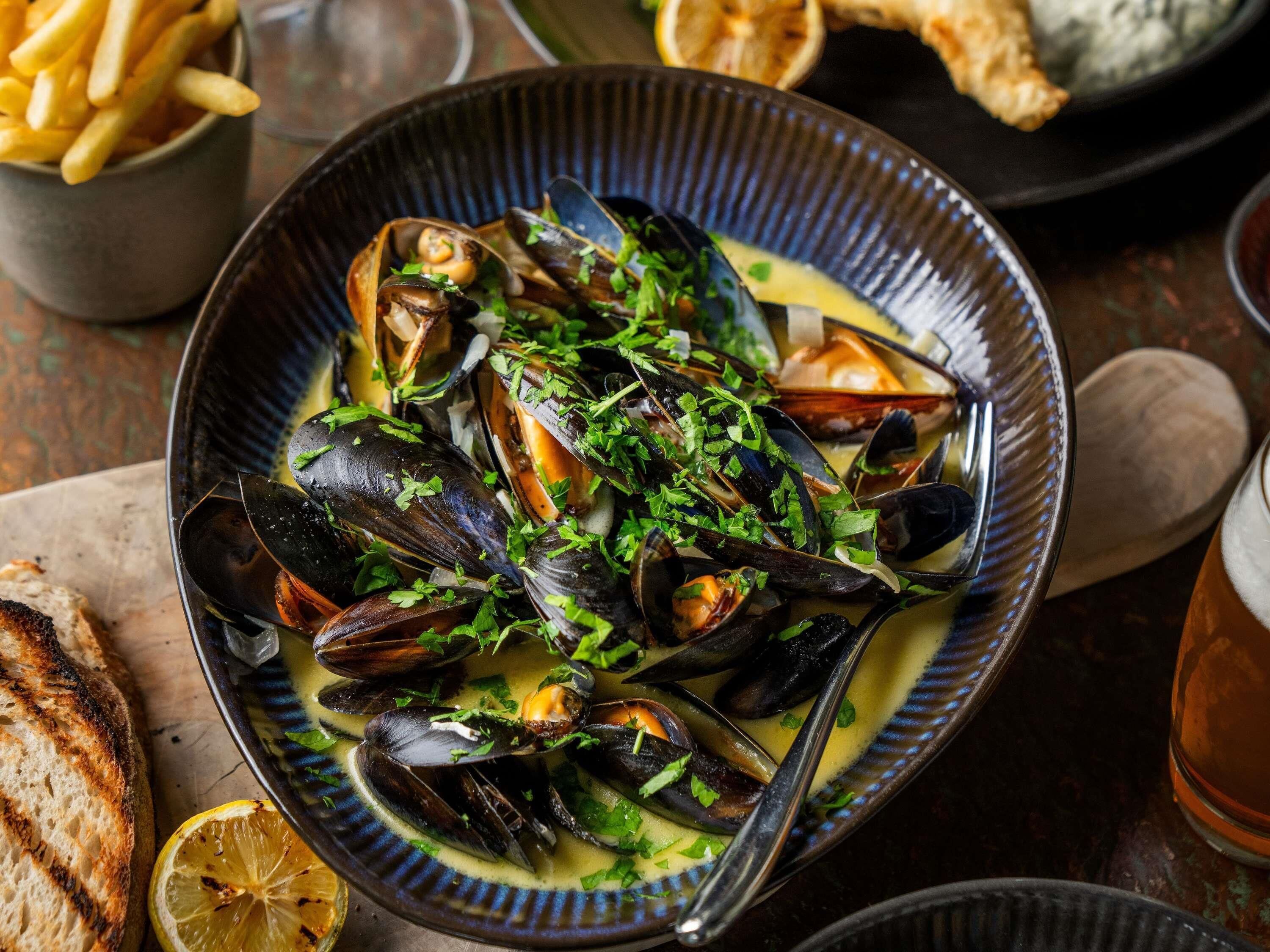 A bowl of mussels garnished with herbs, accompanied by grilled bread and a lemon slice.