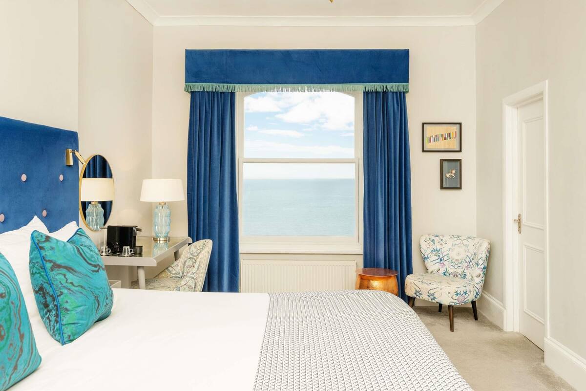 A hotel room with a bed, a desk with a lamp, a chair, a window with curtains, and a view of the sea.