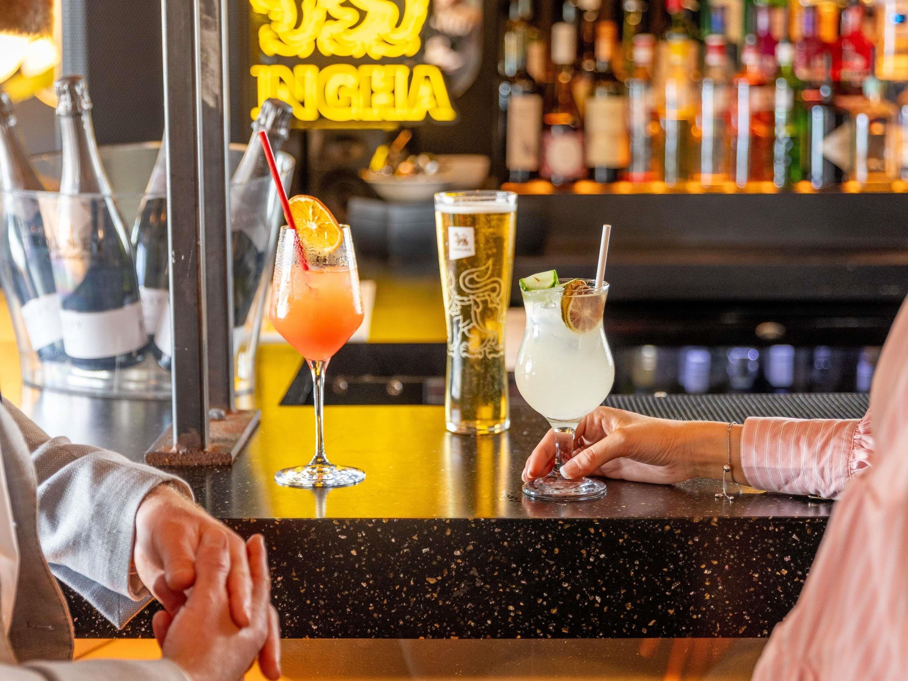 A bar counter with three drinks, a neon sign, and a variety of bottles in the background.
