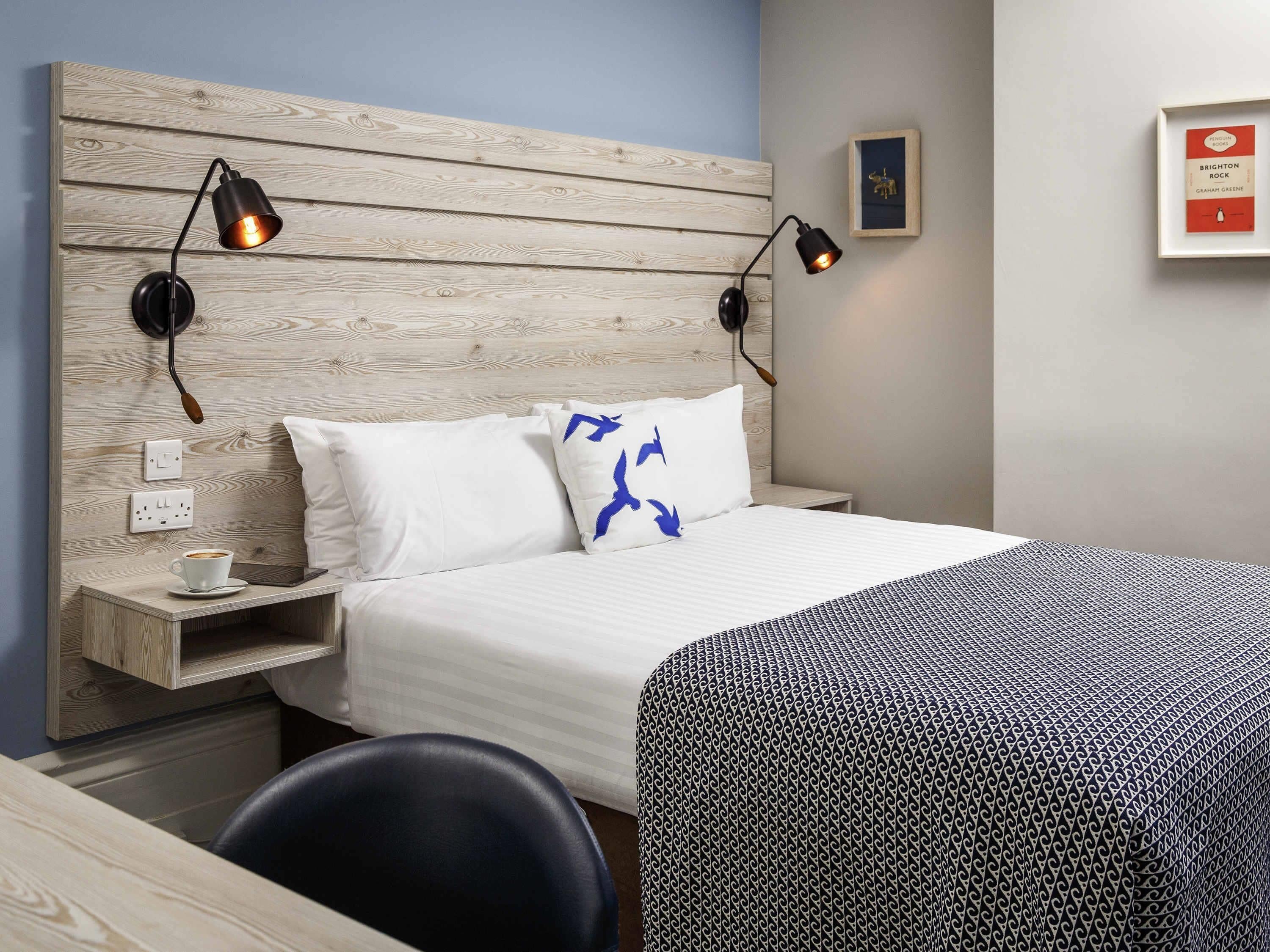 A hotel room with a wooden headboard, two bedside lamps, a bed with white bedding and a blue pillow, a bedside table with a cup and a book, and a framed picture on the wall.