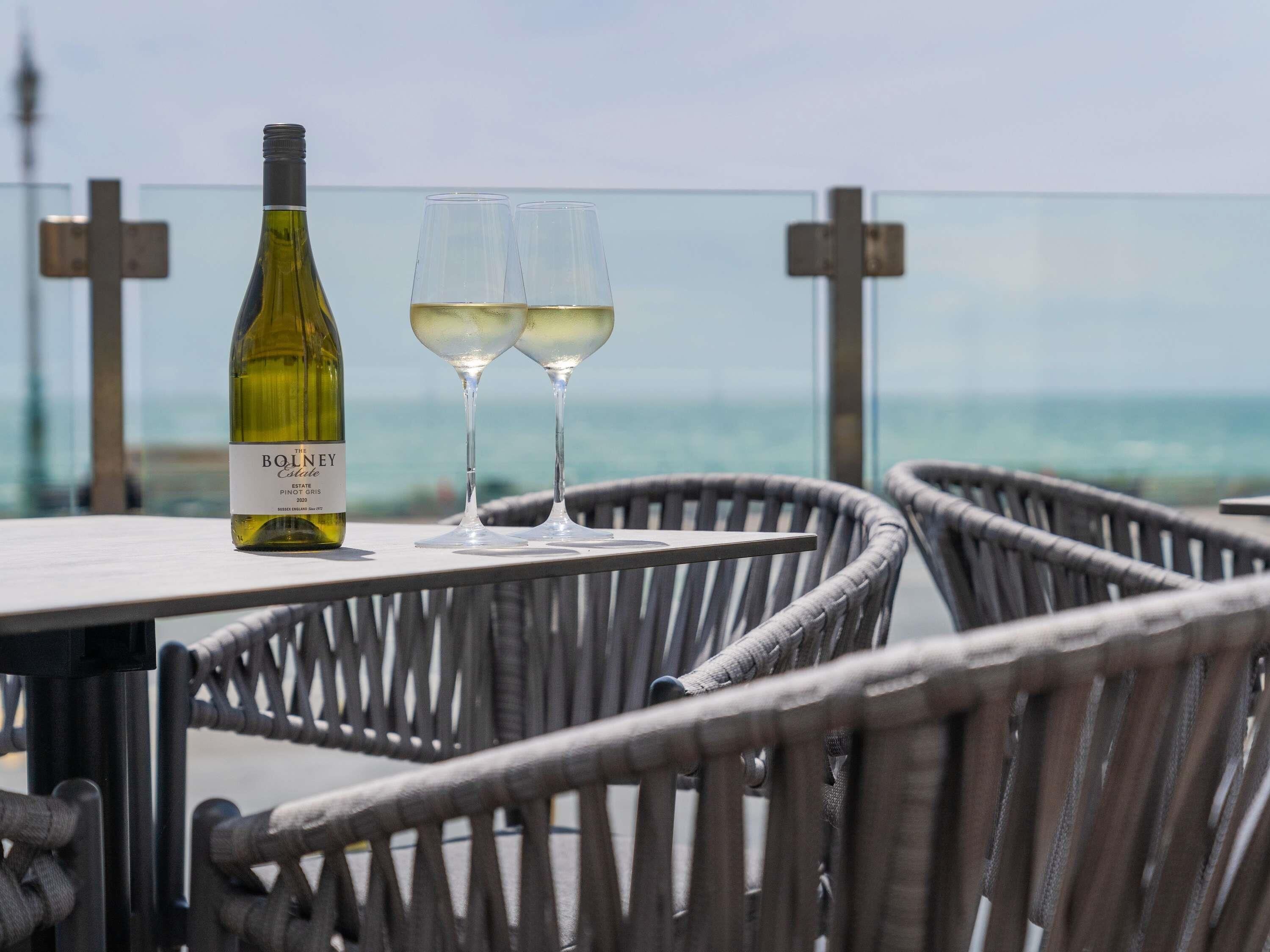 A bottle of Bolney Estate Pinot Grigio wine and two glasses on a table with wicker chairs, overlooking the sea.