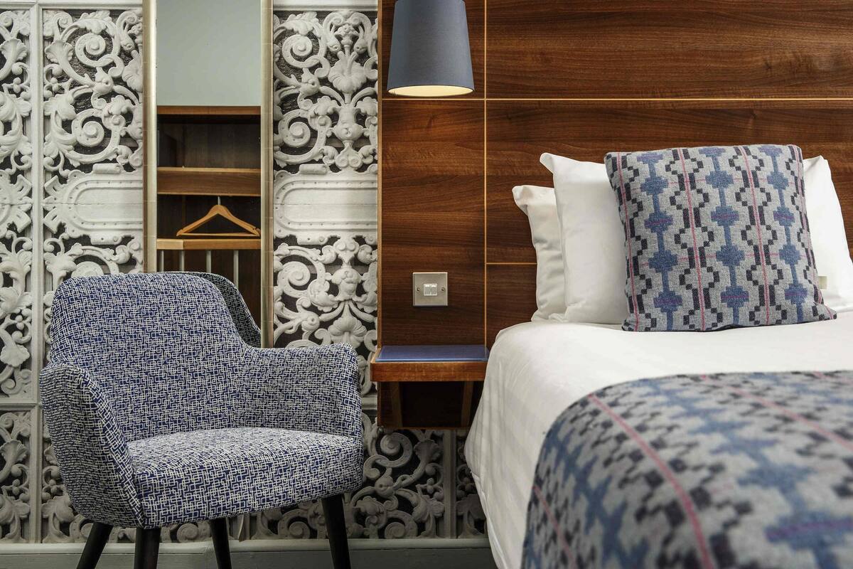 A hotel room with a patterned wall, a wooden headboard, a bedside lamp, a patterned bedspread, a patterned pillow, and a patterned chair.