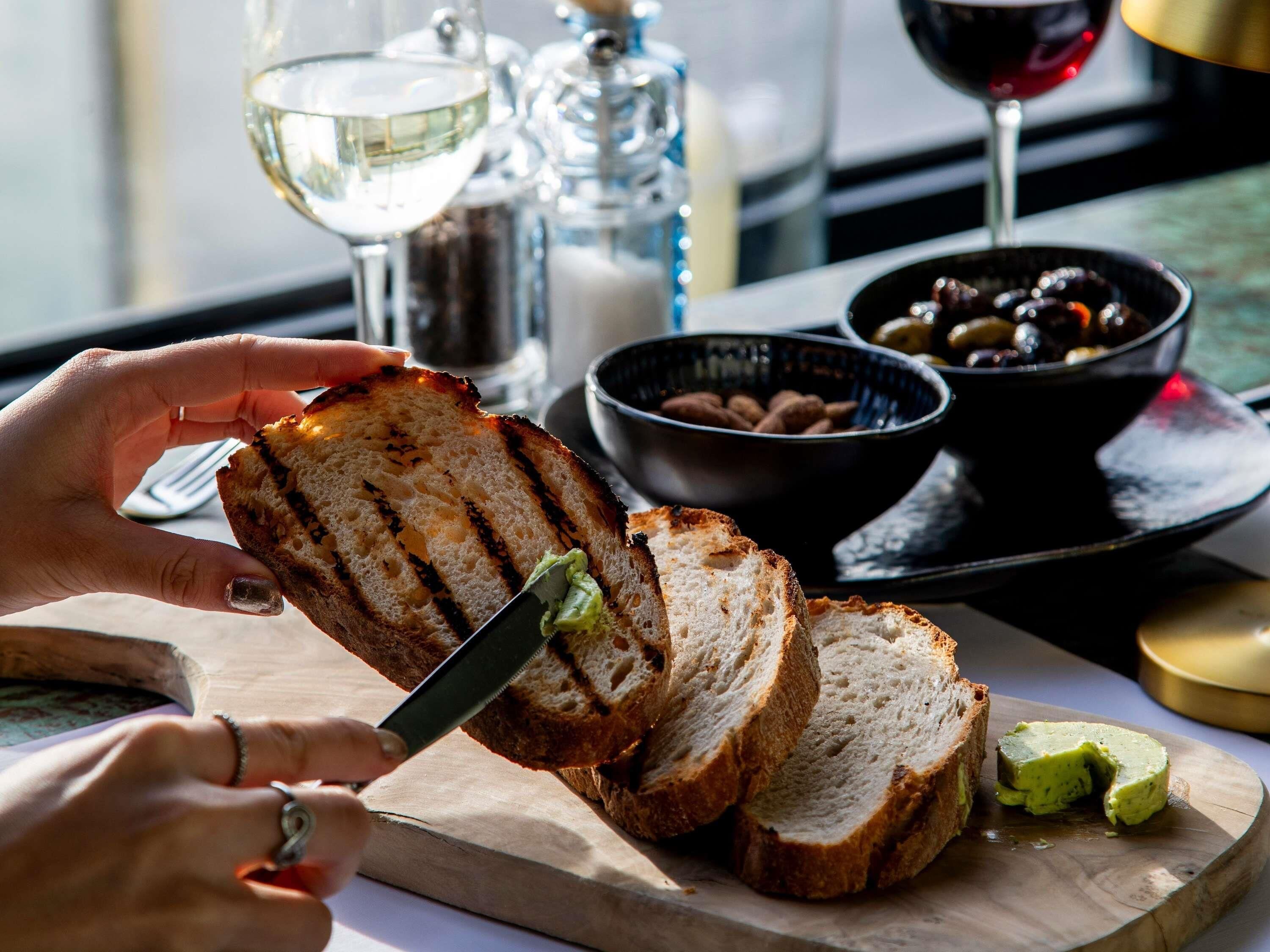 Une personne coupe du pain grillé sur une planche en bois à l’aide d’un couteau, et à côté, on voit un bol d’olives et un verre de vin blanc.