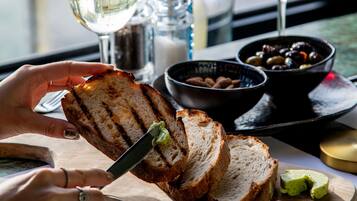 Une personne coupe du pain grillé sur une planche en bois à l’aide d’un couteau, et à côté, on voit un bol d’olives et un verre de vin blanc.