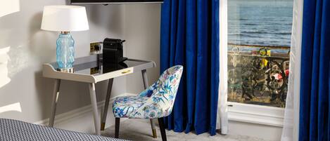 A hotel room with a bed, a desk with a lamp, a chair, a TV, and a window with a view of the sea.