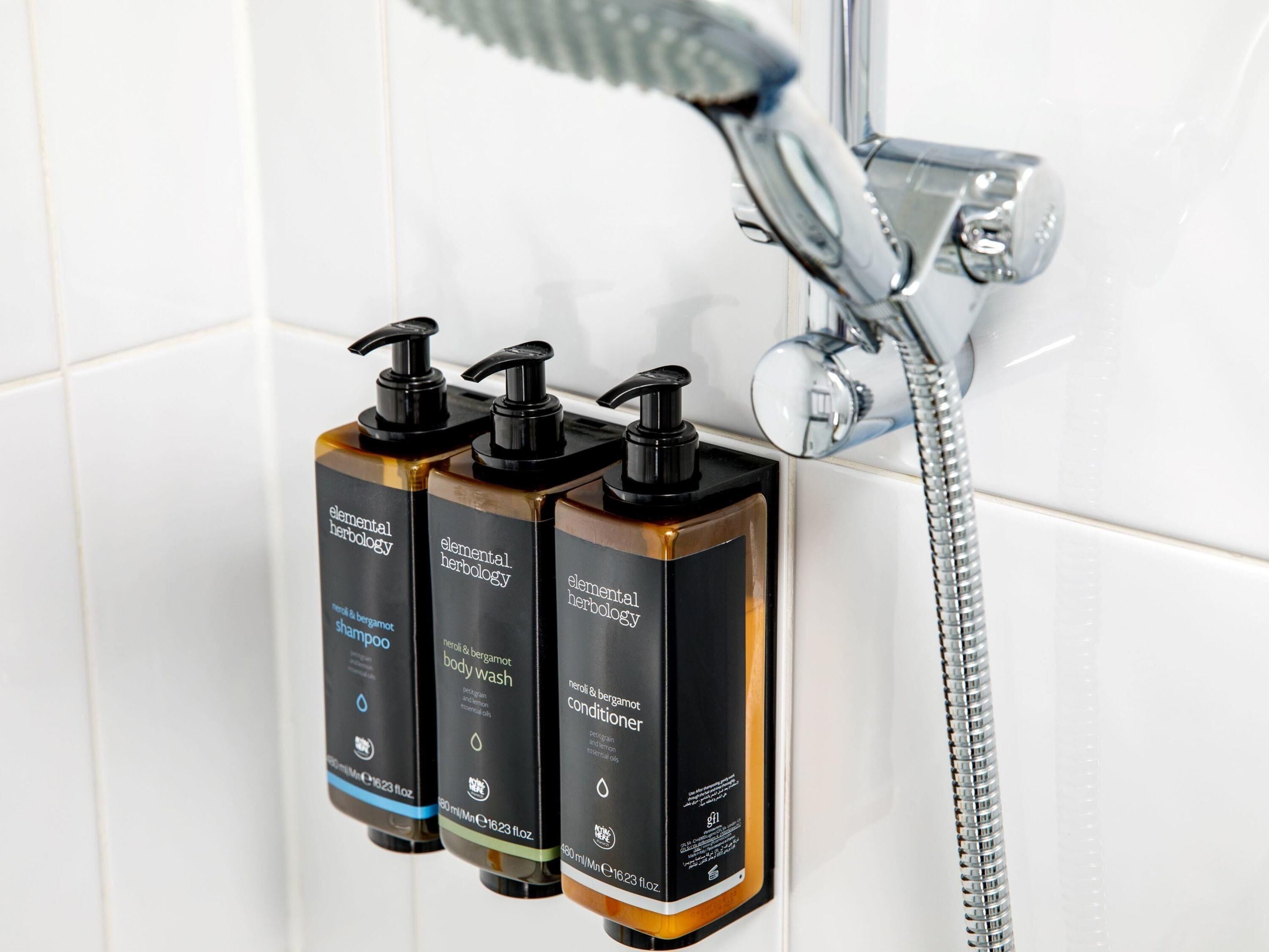 Three bottles of Elemental Herbology shampoo, body wash, and conditioner mounted on a wall near a shower.