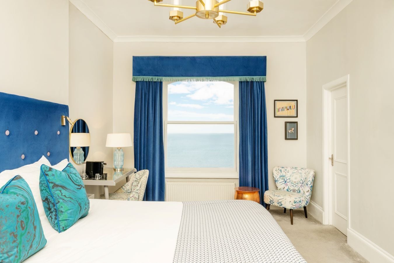 Ein Schlafzimmer mit einem großen Bett, einem Schreibtisch mit Lampe, einem Stuhl und Blick aufs Meer, sowie einer Tür.
