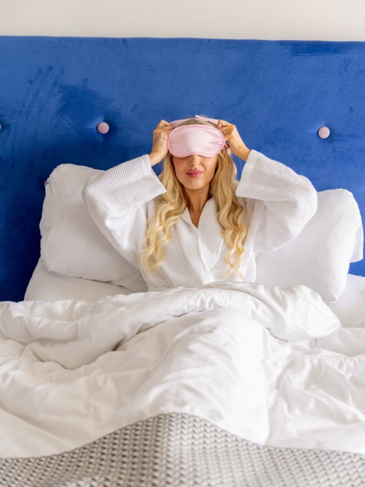 A person in a white robe sitting on a bed with white bedding, a blue headboard, and two round wall-mounted lights.