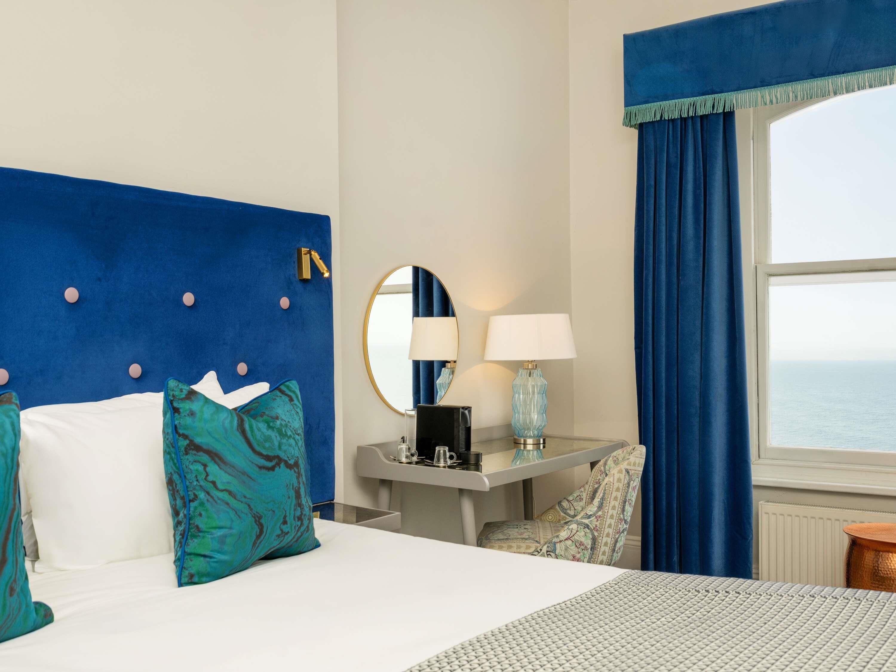A hotel room with a bed, a desk with a lamp, a chair, a round mirror, and a view of the sea.