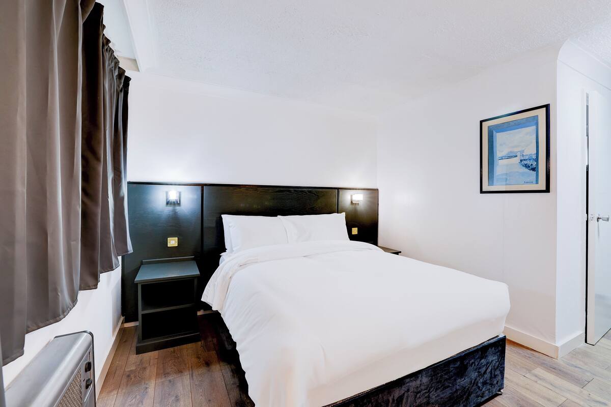 A bedroom with a bed, a nightstand, a wall-mounted light, and a framed picture.