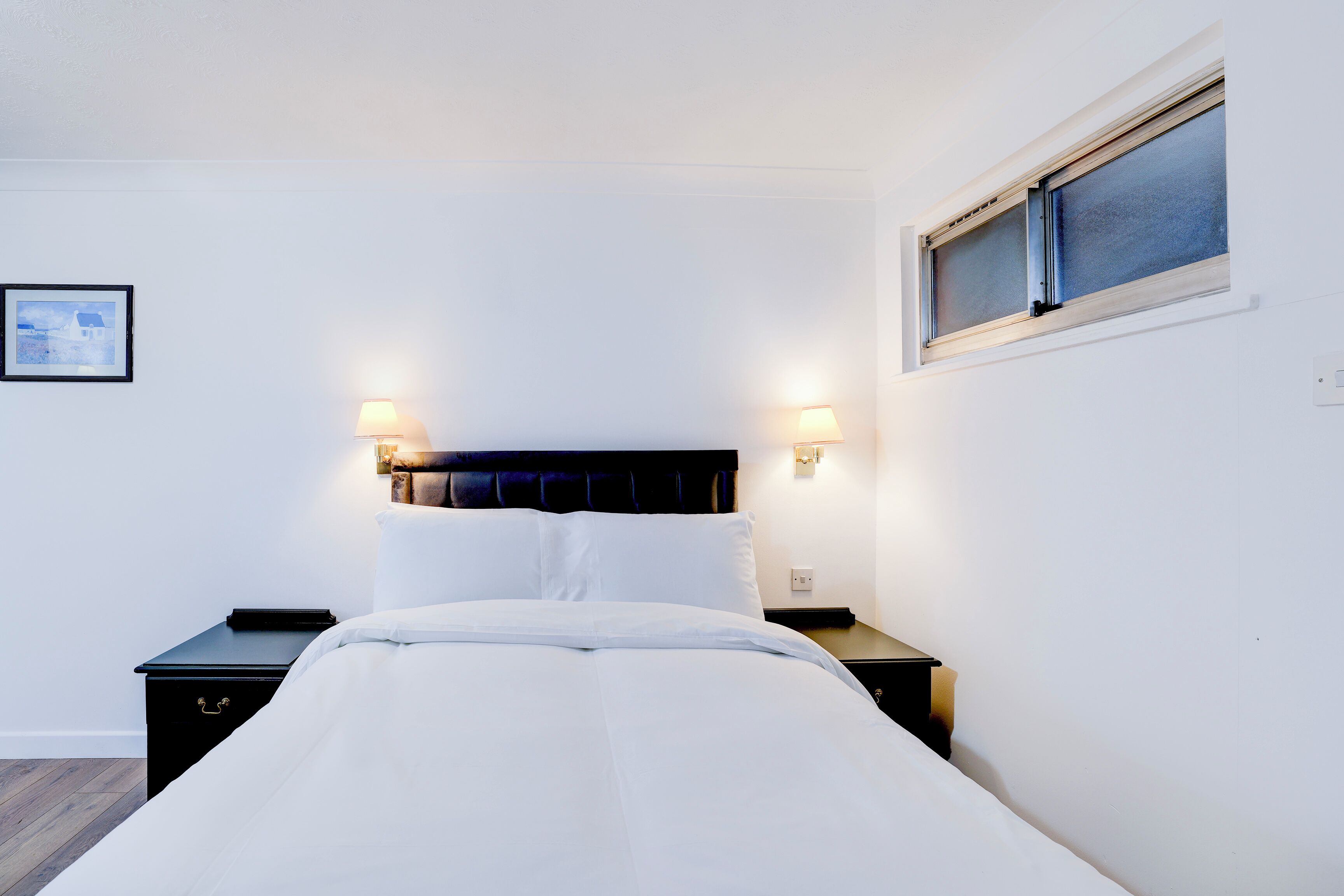 A neatly made bed with white linens, two bedside tables with lamps, and a window with blinds.