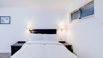 Una cama bien hecha con sábanas blancas, dos mesitas de noche con lámparas y una ventana con persianas.