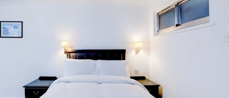 A neatly made bed with white linens, two bedside tables with lamps, and a window with blinds.