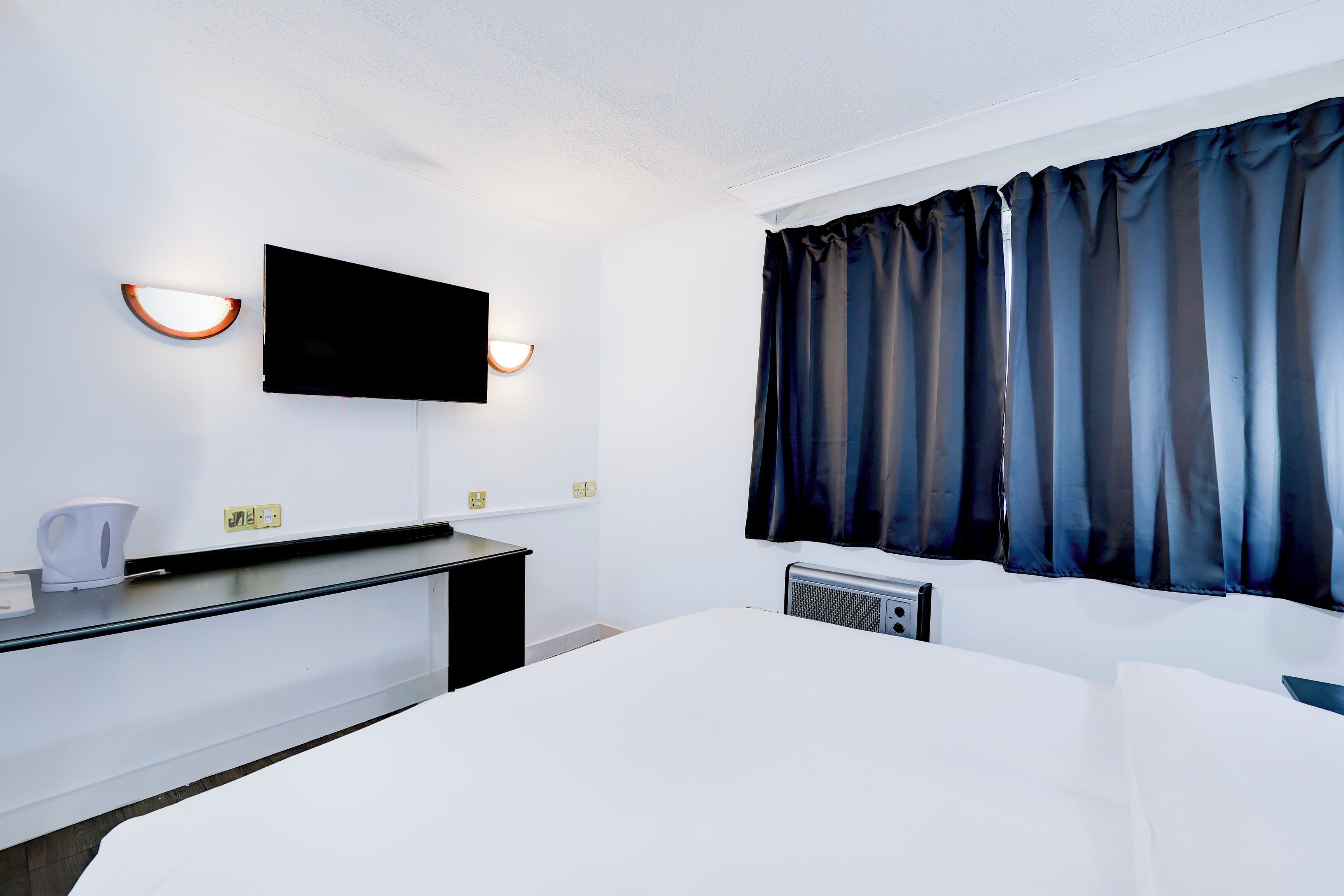 Quarto de hotel com cama, televisão de tela plana na parede, escrivaninha com chaleira e janela com cortinas escuras.