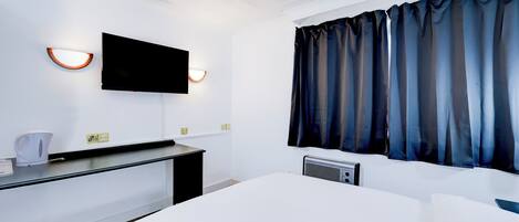 Quarto de hotel com cama, televisão de tela plana na parede, escrivaninha com chaleira e janela com cortinas escuras.