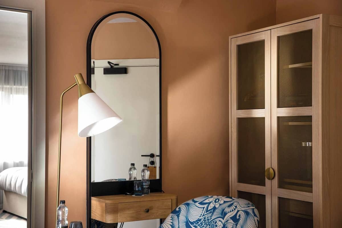 Een slaapkamer met een bed, een bedlampje, een spiegel, een houten kast en een dressoir met toiletartikelen.