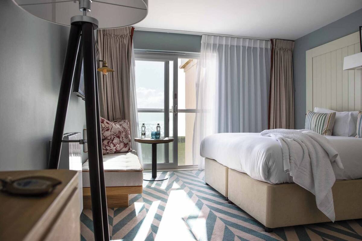 Hotelkamer met een bed, een stoel, een tafel met flessen, een lamp en uitzicht door een glazen deur.