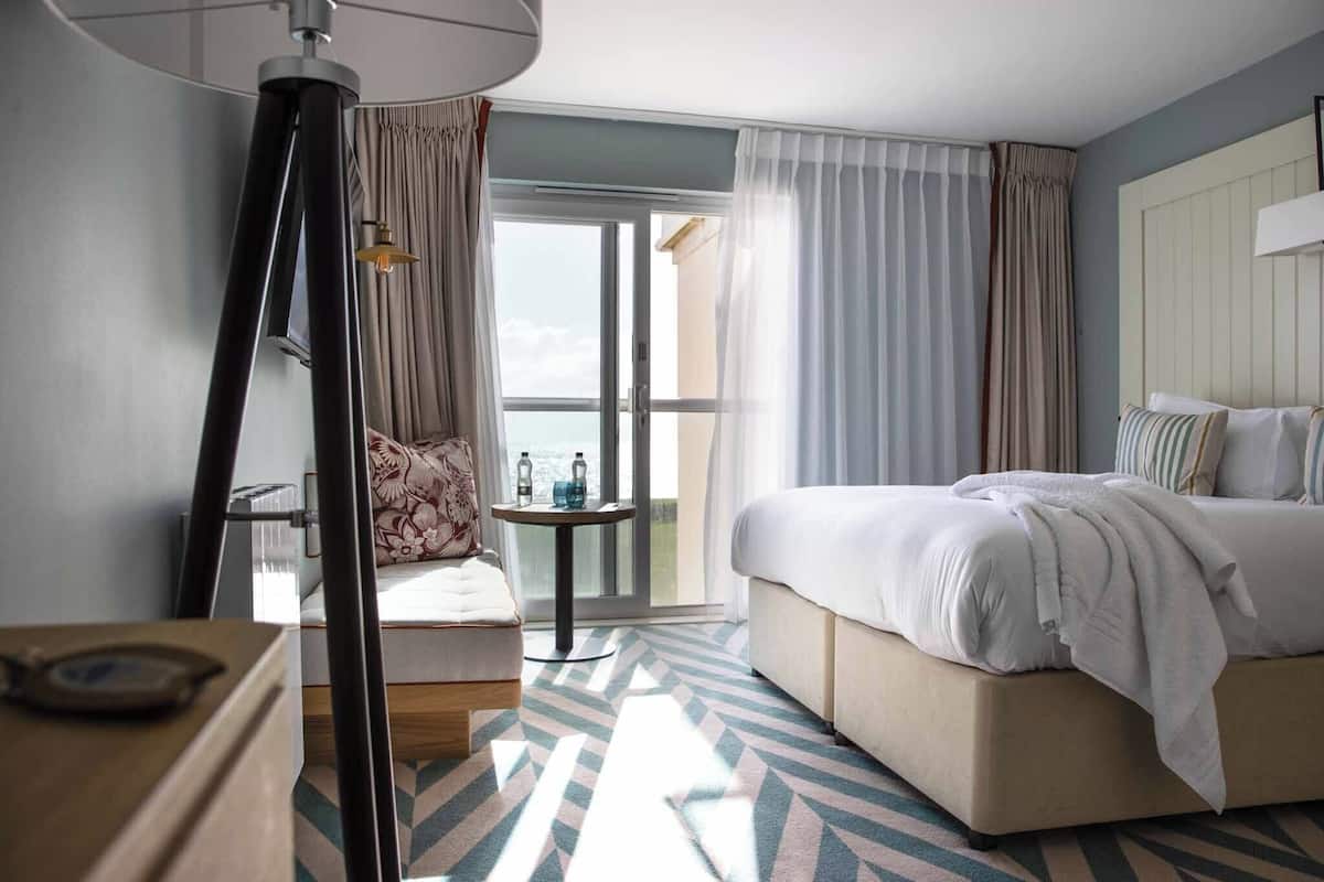 Ein Hotelzimmer mit Bett, Sessel, Tisch mit Flaschen, Lampe und Blick durch eine Glastür.