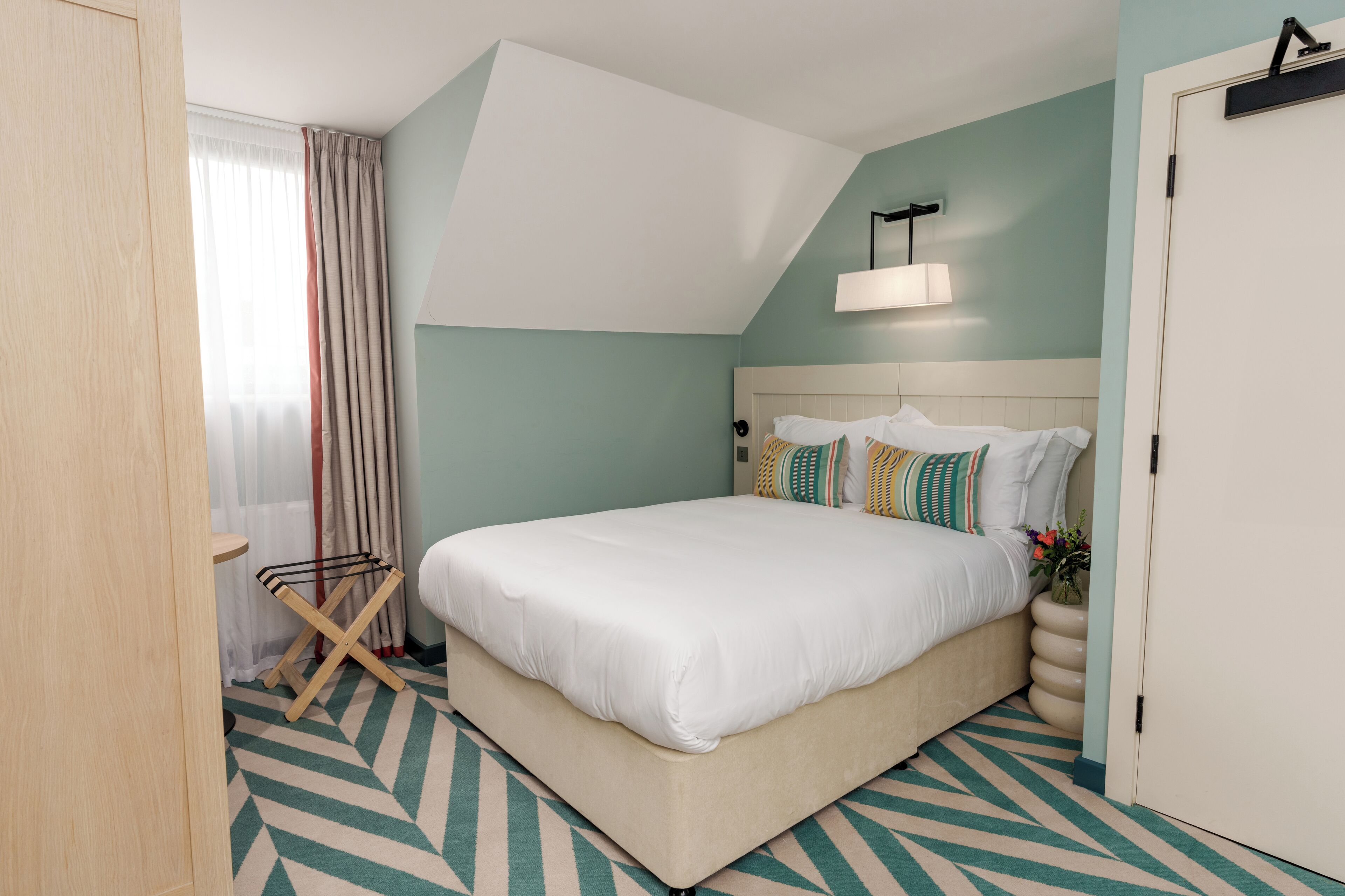 A neatly arranged bedroom with a large bed, a bedside table, a lamp, and a window with curtains.