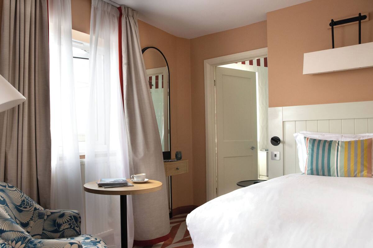 Een hotelkamer met een bed, een stoel, een klein tafeltje met een kopje en gordijnen.
