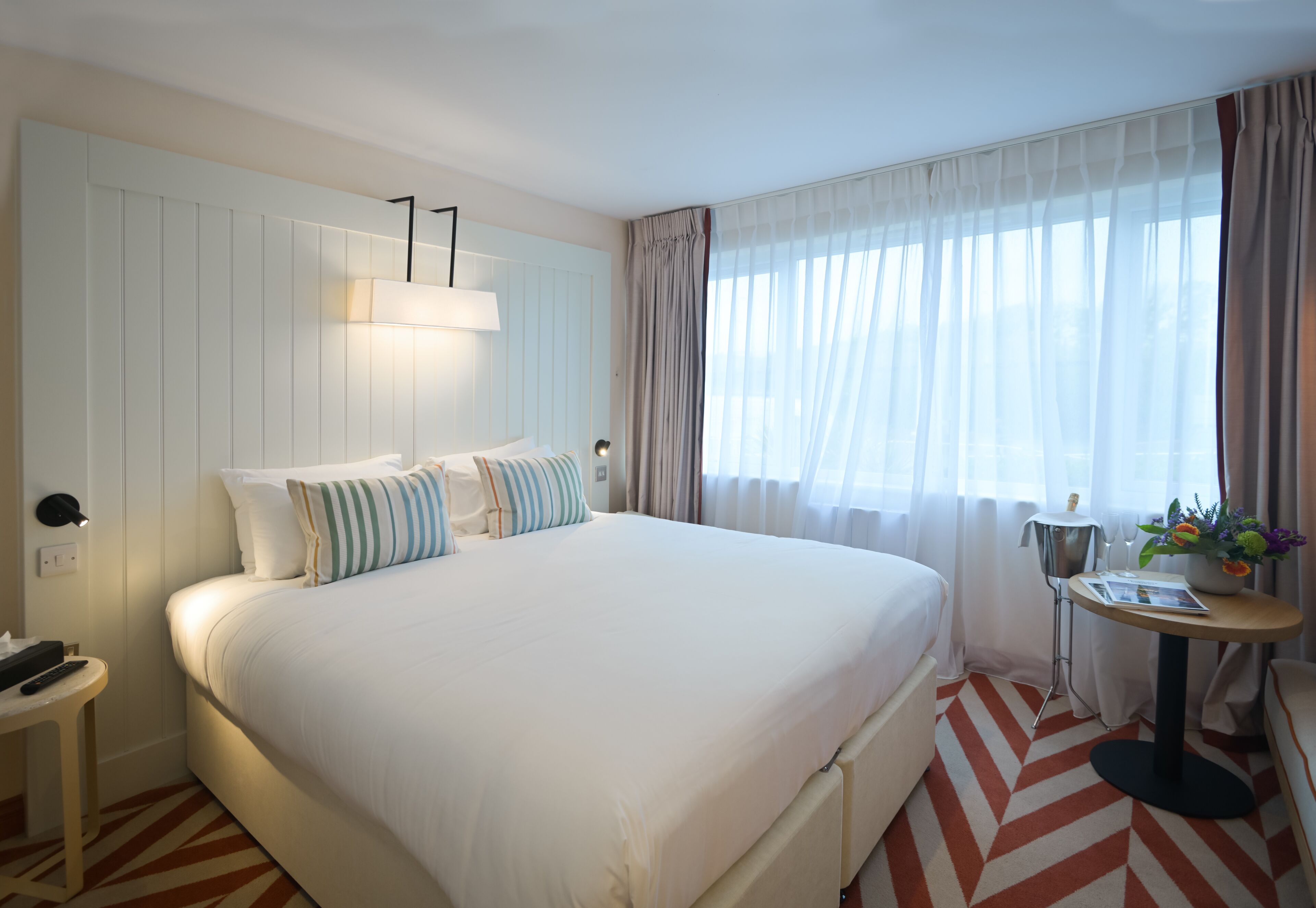 Una camera d'albergo con un letto grande, un comodino con una lampada, un tavolino con un vaso di fiori e una finestra con tende leggere.