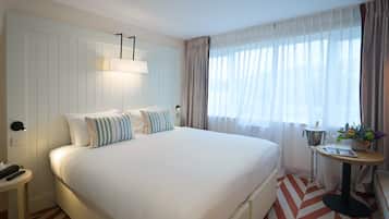 Una camera d'albergo con un letto grande, un comodino con una lampada, un tavolino con un vaso di fiori e una finestra con tende leggere.