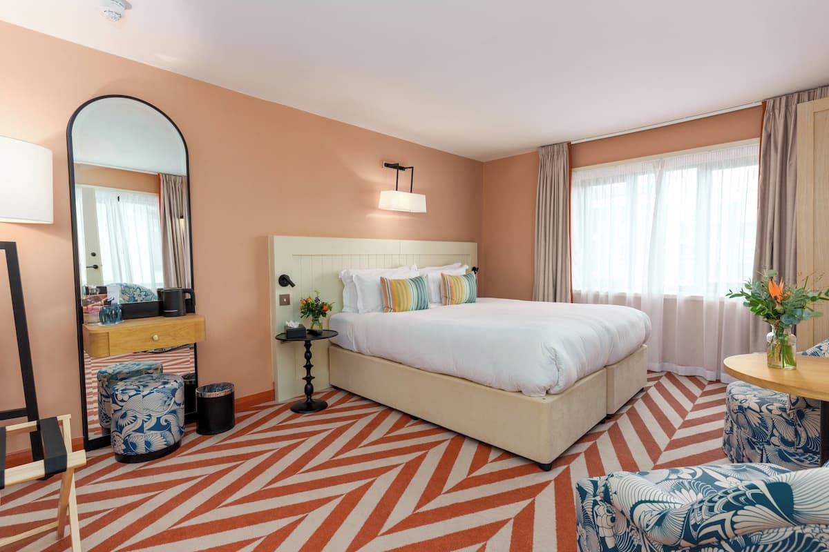 Een hotelkamer met een groot bed, een nachtkastje met een lamp, een spiegel en een kamerkleed met een patroon.