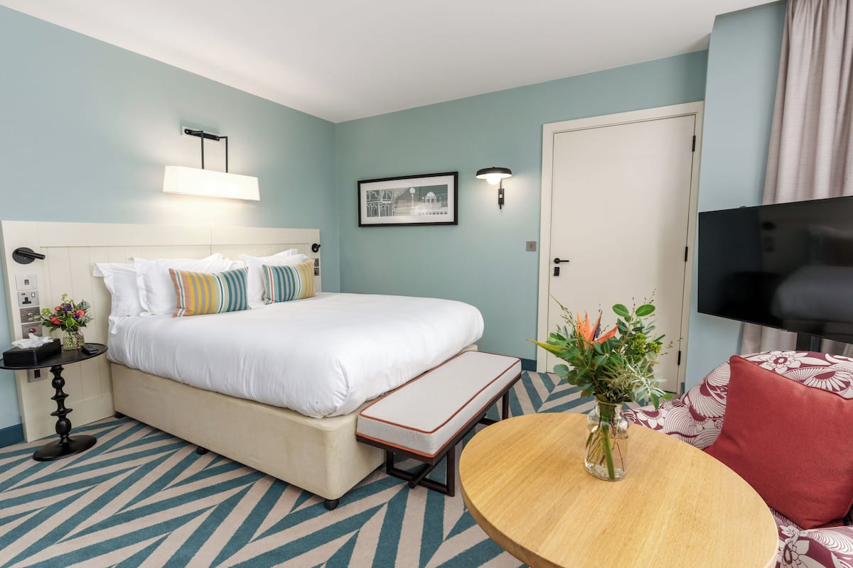 Een hotelkamer met een bed, nachtkastje, lamp, televisie en een klein rond tafeltje met een bloevas.