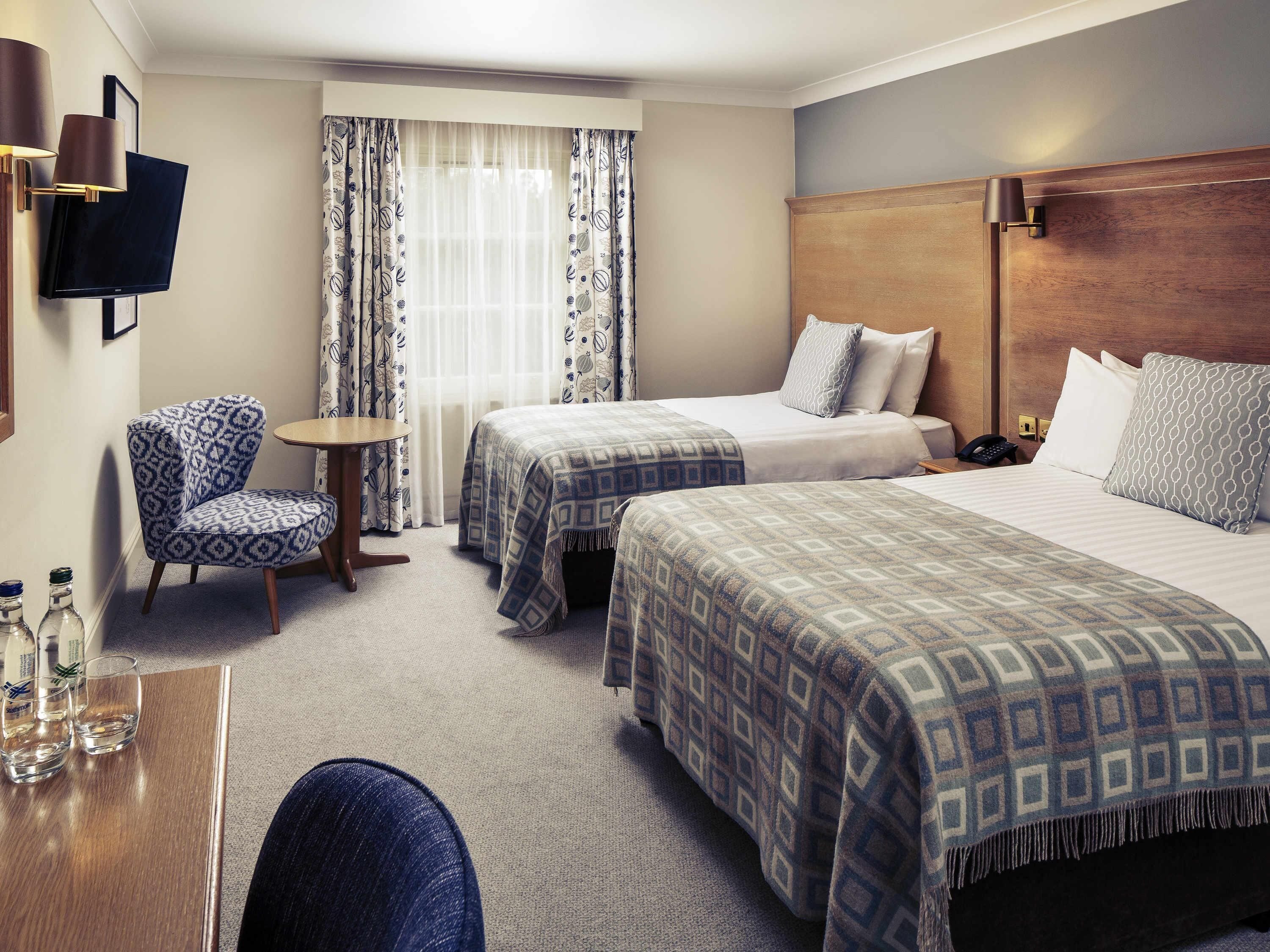 A hotel room with two beds, a chair, a small table, a TV, and a lamp.