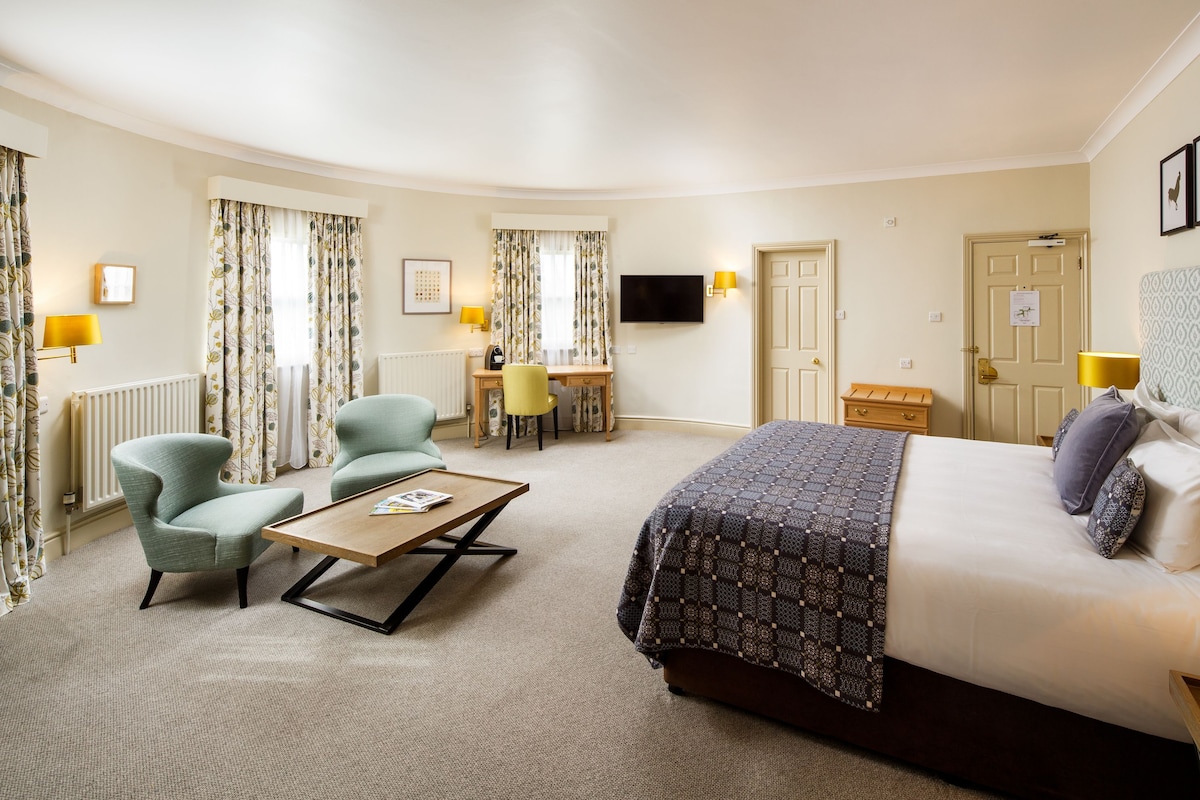 A hotel room with a large bed, a desk, two chairs, a TV, and a window with curtains.