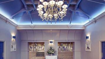 A grand hall with a large chandelier, a staircase leading to a banquet area, and framed artwork on the walls.