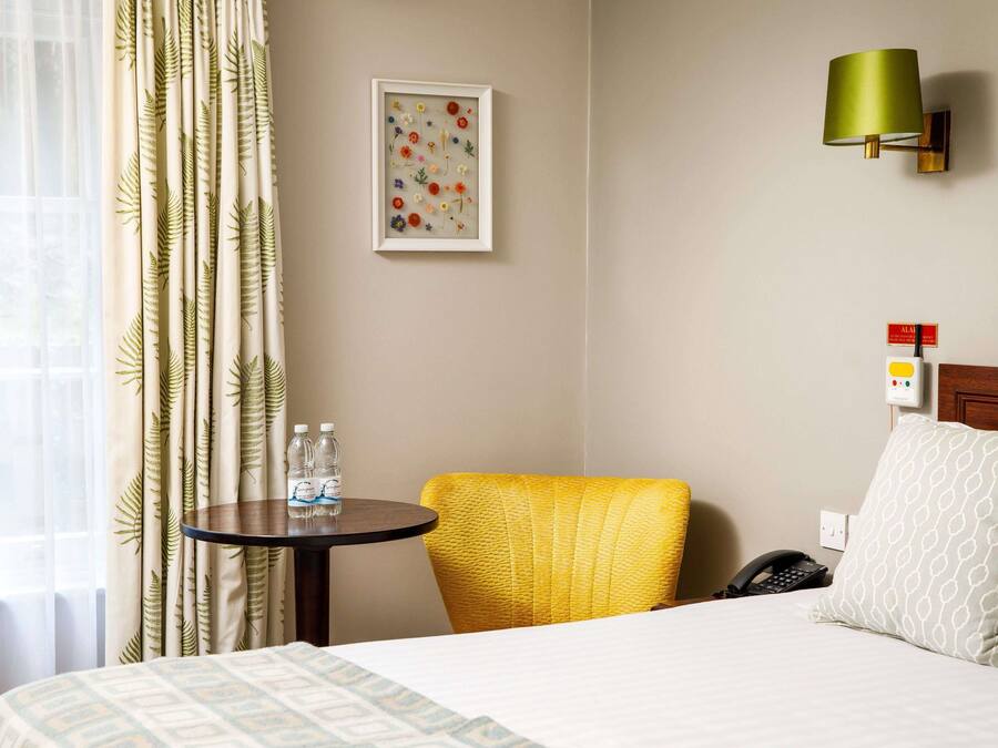 A hotel room with a bed, a small table with water bottles, a yellow chair, a framed floral artwork, a wall-mounted lamp, and a bedside phone.