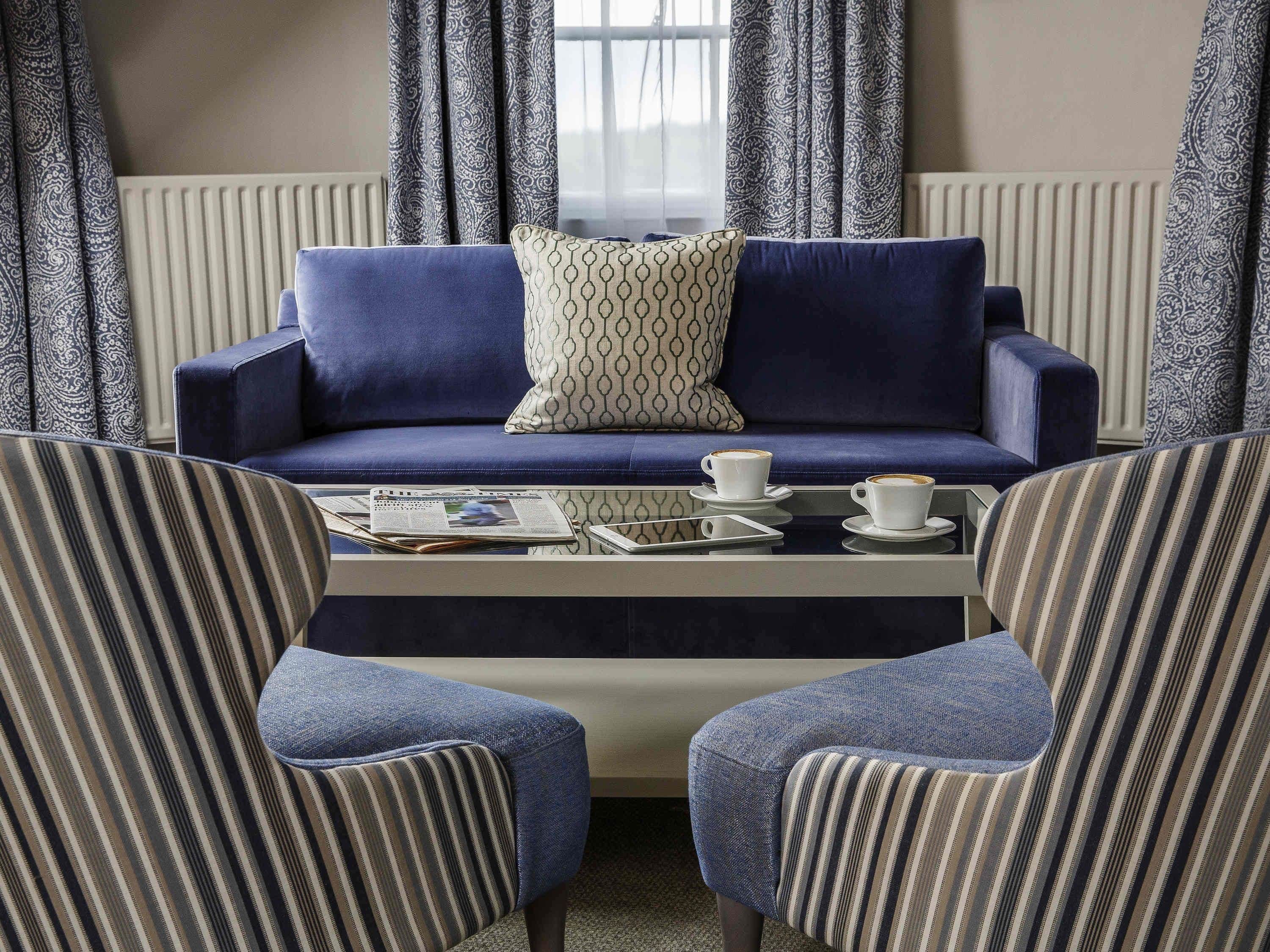 A modern living room with a blue sofa, patterned cushions, a coffee table with newspapers and a smartphone, and striped armchairs.
