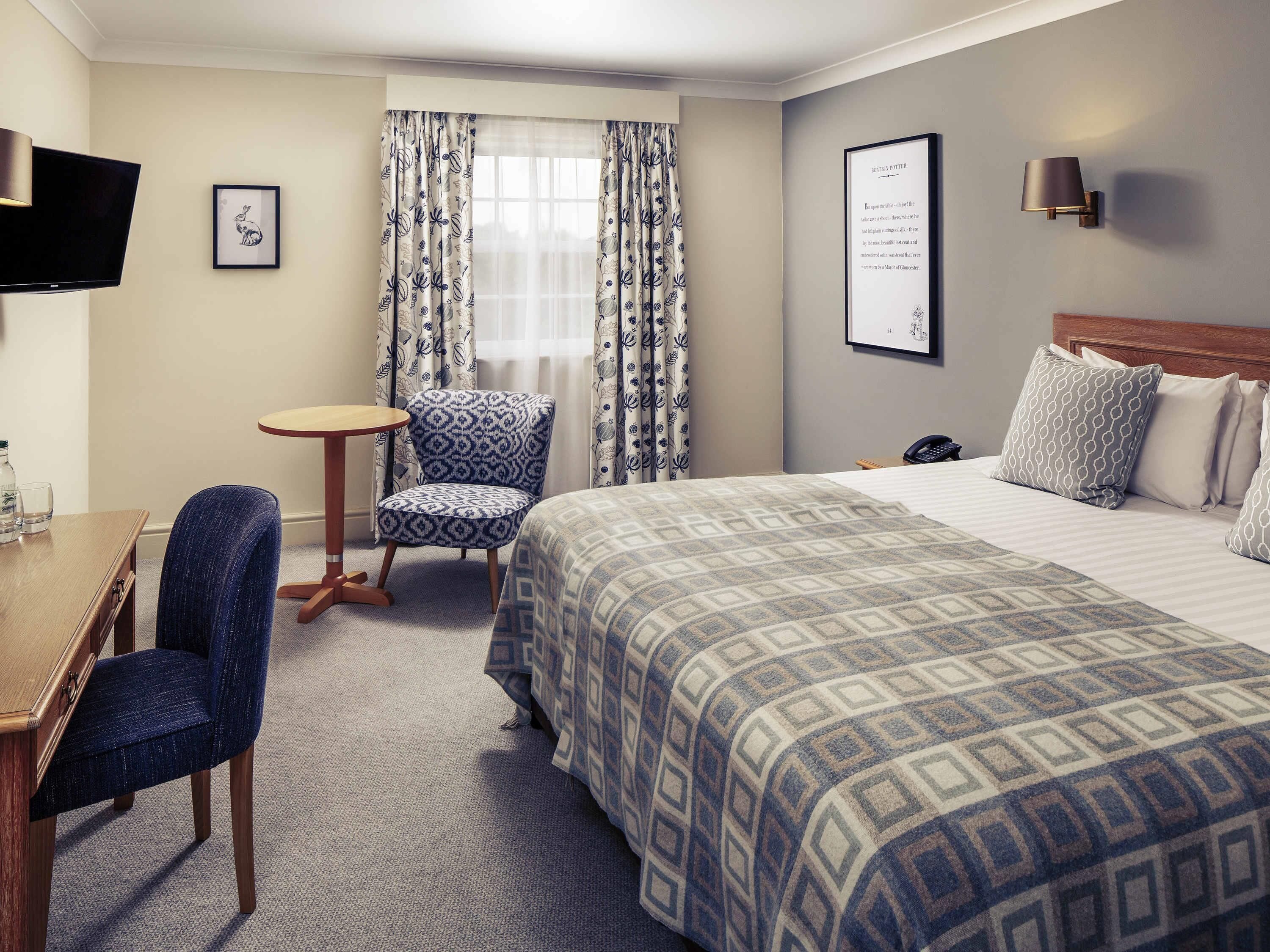 A hotel room with a bed, a chair, a small table, a TV, and a framed quote on the wall.