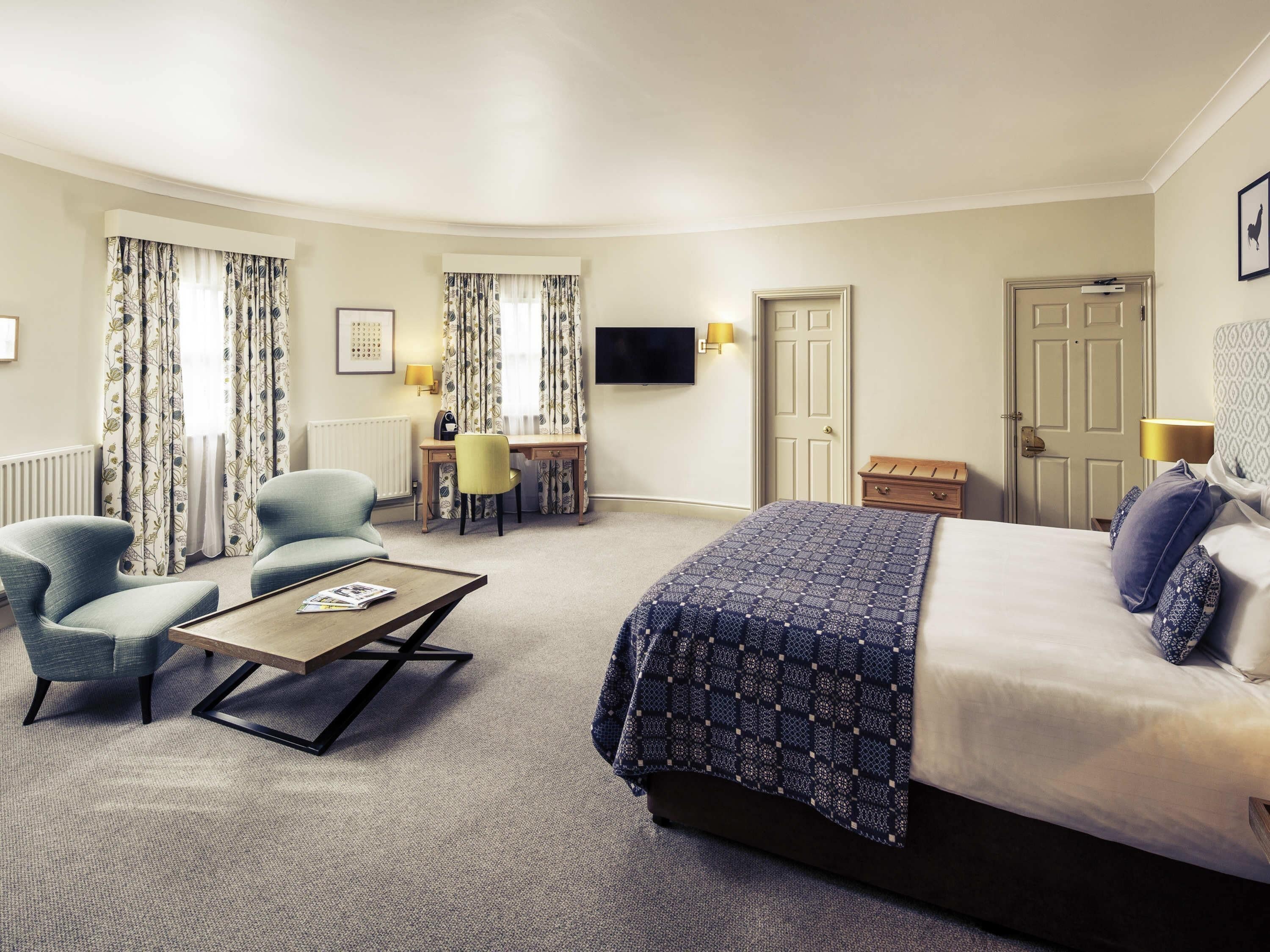 A hotel room with a bed, two armchairs, a desk, a television, and a door.