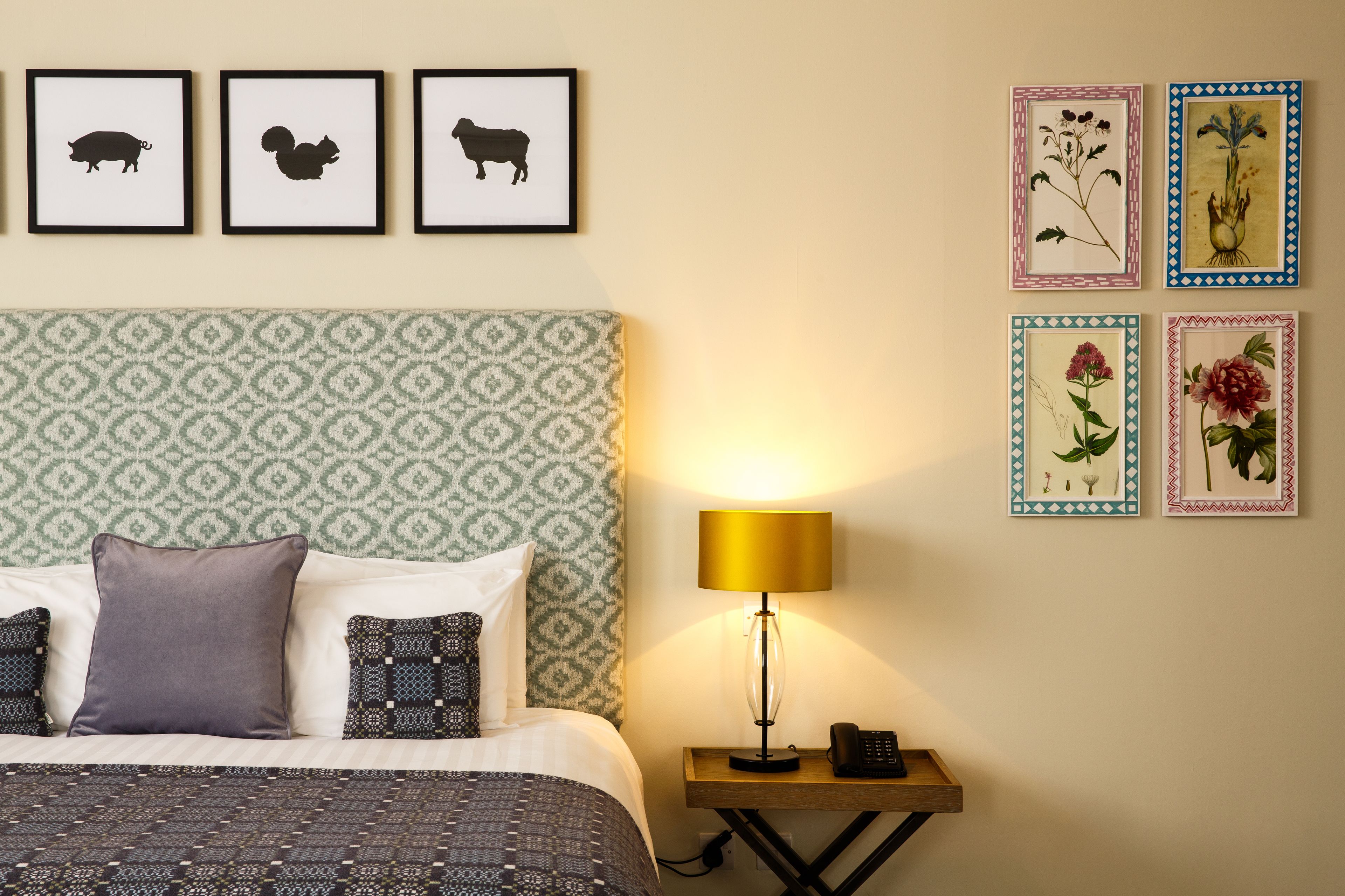 Una cama bien hecha con ropa de cama estampada, una lámpara de noche e impresiones enmarcadas de animales y plantas en la pared.