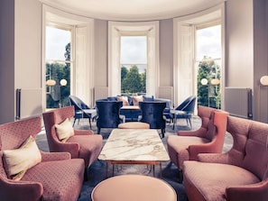 A modern room with a marble coffee table, plush seating, and large windows offering a view of greenery. - Mercure Gloucester Bowden Hall Hotel (Gloucester)