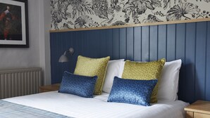 A neatly made bed with patterned pillows, a blue headboard, and a floral wallpaper design. - The Bear Hotel by Greene King Inns (Hungerford)