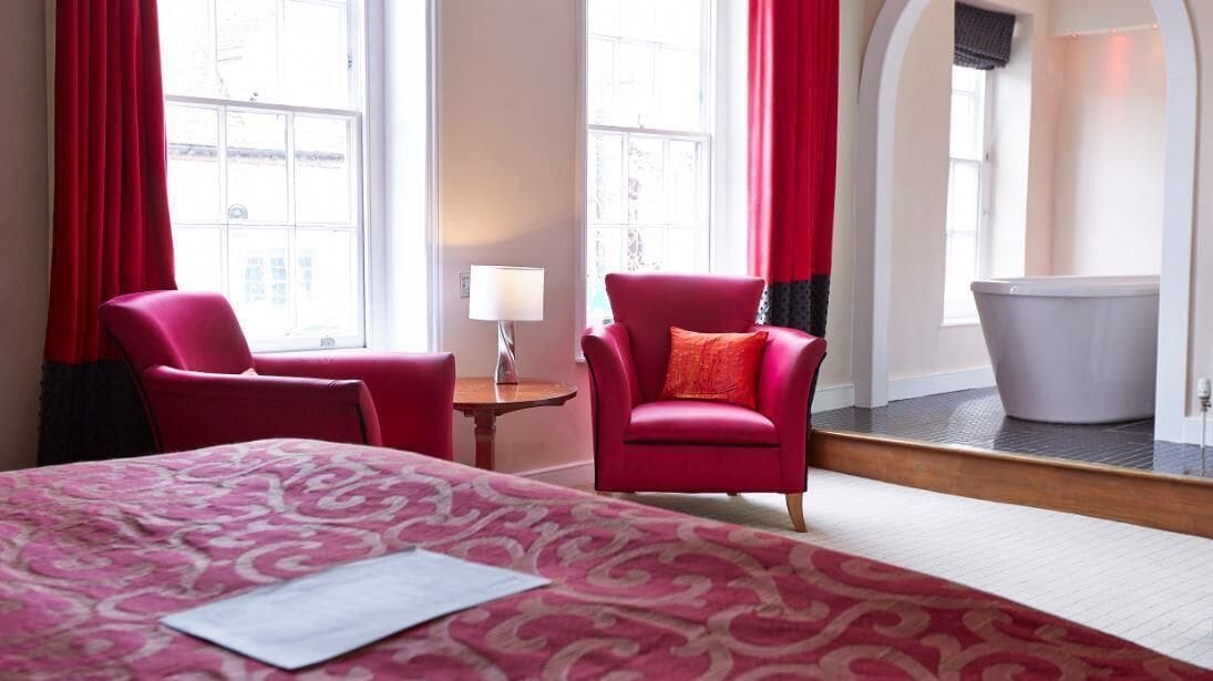 Une chambre d’hôtel avec un grand lit, deux fauteuils rouges, une petite table avec une lampe et une baignoire.