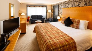 Ein Hotelzimmer mit einem großen Bett, einem Fernseher, einem Schreibtisch mit Lampe, einer Couch und einem Sessel.