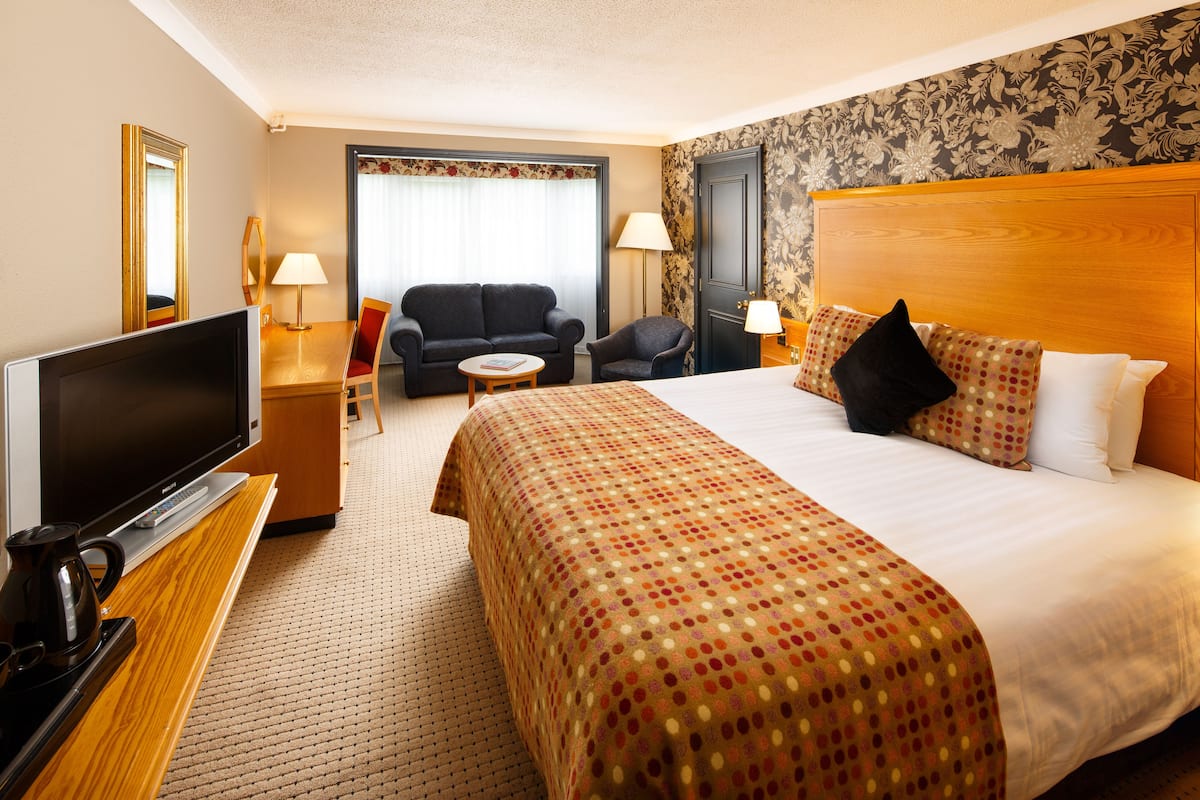 A hotel room with a large bed, a television, a desk with a lamp, a sofa, and a chair.