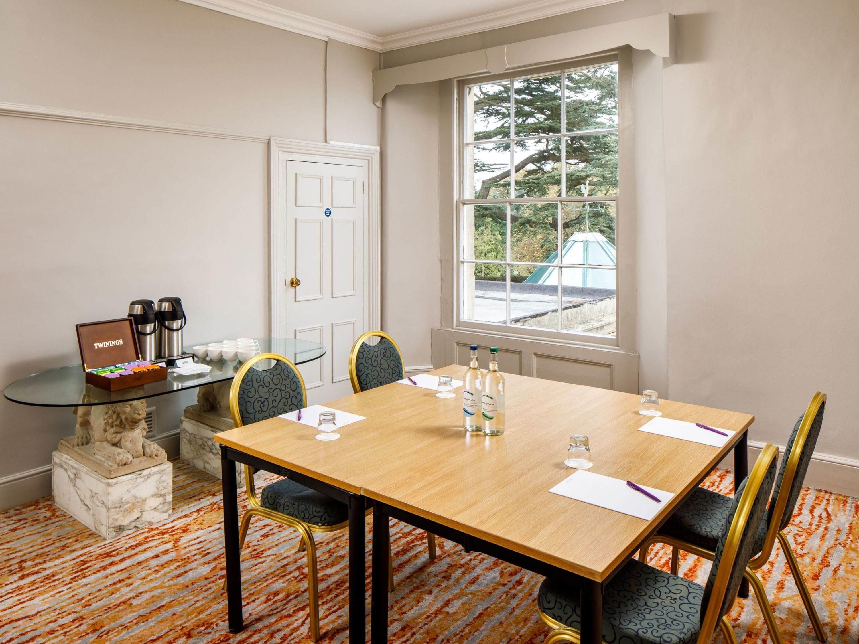 A conference room with a wooden table, chairs, a glass coffee table, a statue, and a window with a view of greenery.