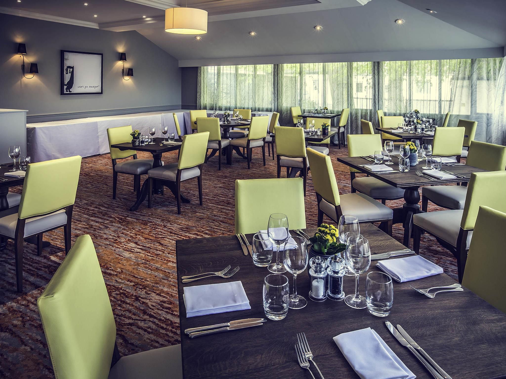 A hotel dining area with multiple tables set for guests, featuring a carpeted floor and natural light from large windows.