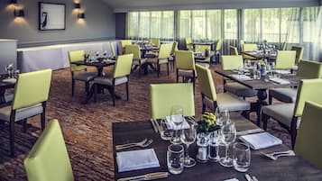 A hotel dining area with multiple tables set for guests, featuring a carpeted floor and natural light from large windows.
