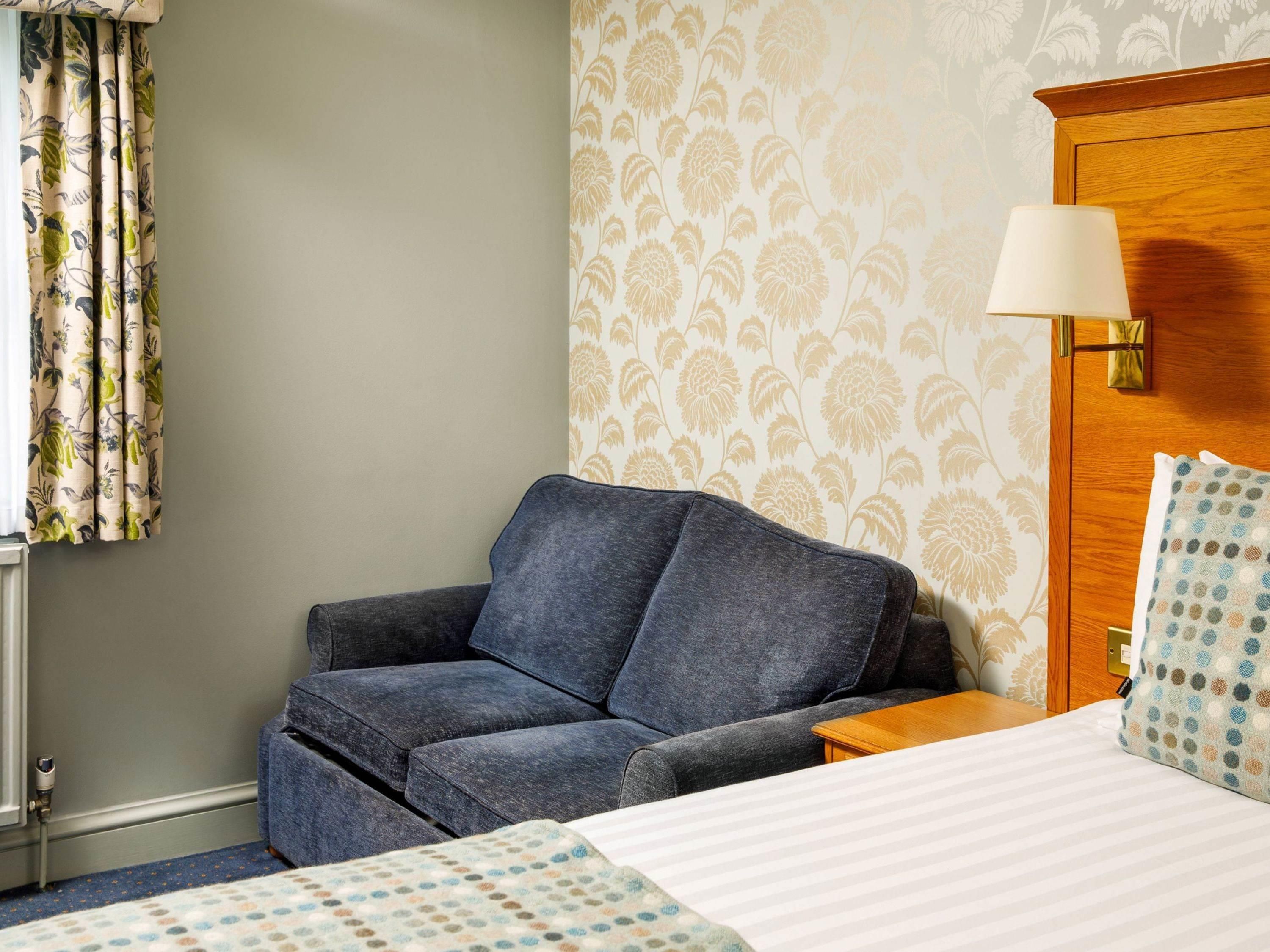 A hotel room with a grey sofa, a wooden nightstand, a bed with a striped bedspread, a wall with floral wallpaper, and a window with curtains.