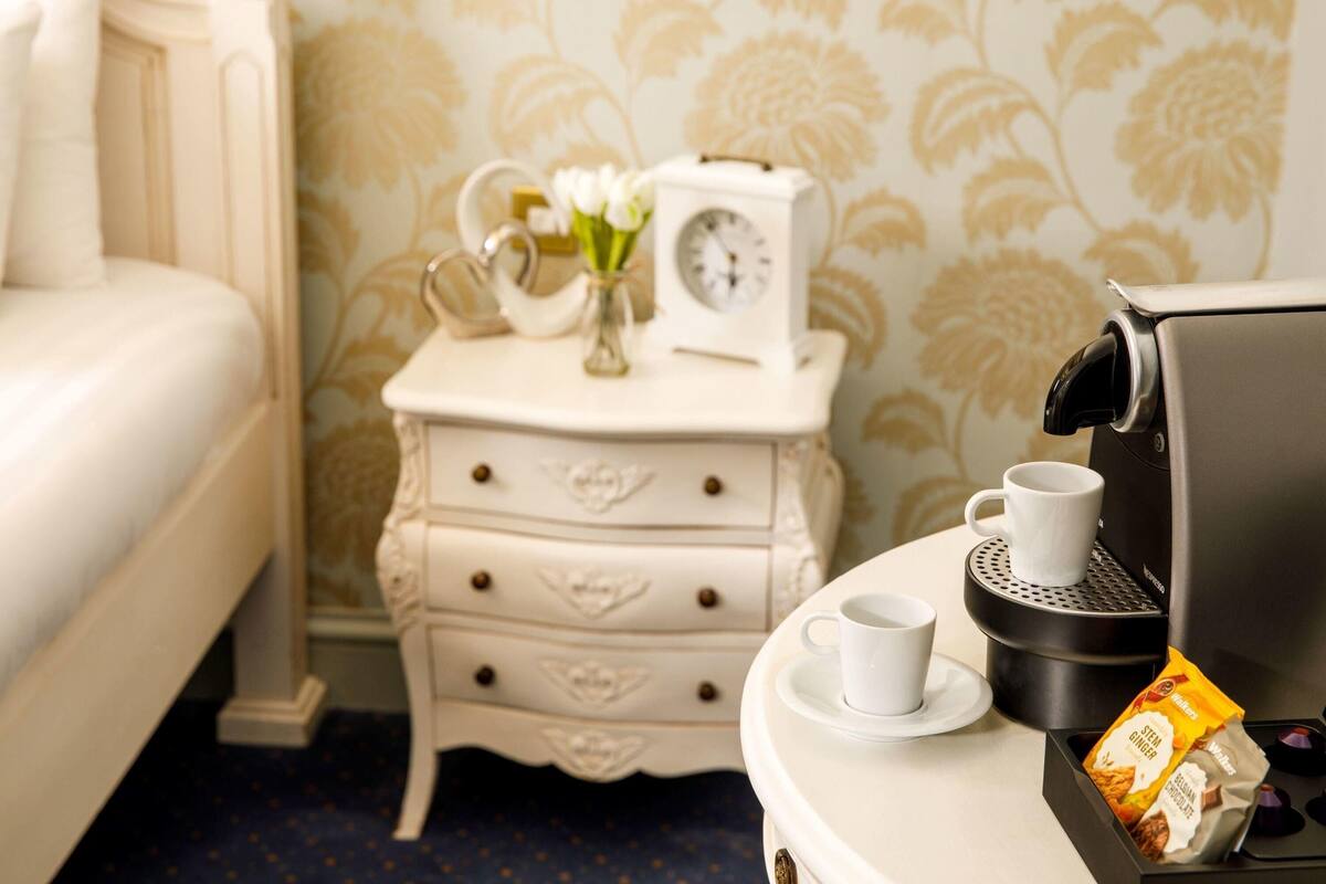 A neatly made bed, a bedside table with a clock and a vase of flowers, a coffee machine with a cup and a packet of coffee, and a patterned wall.