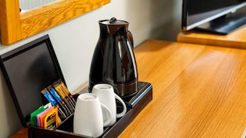 Een hotelkamer met een houten bijzettafeltje waarop een koffiezetapparaat, twee witte mokken en een dienblad met verschillende koffiehoezen van verschillende merken staan.