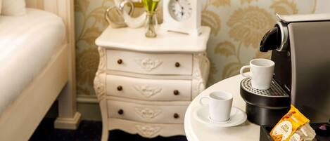 Un lit bien fait, une table de chevet avec une horloge et un vase de fleurs, une machine à café avec une tasse et un paquet de café, et un mur orné d’un motif.